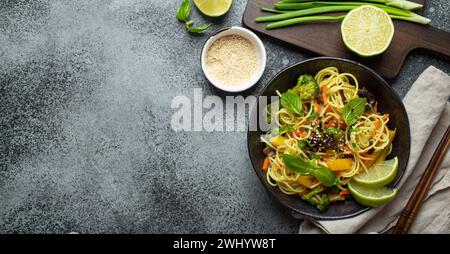 Asian vegetarian noodles with vegetables and lime in black rustic ceramic bowl, wooden chopsticks, cutting board with chopped gr Stock Photo