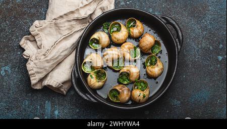 Escargots de Bourgogne Cooked Snails with Garlic Butter and Parsley in black cast iron pan on rustic stone background from above Stock Photo