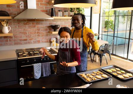 Happy diverse couple in aprons baking christmas cookies, dancing in kitchen, copy space Stock Photo