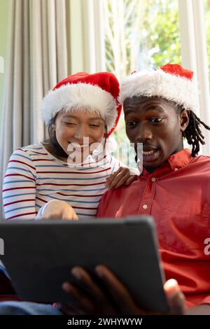 Happy, surprised diverse couple in christmas hats having tablet video call in sunny living room Stock Photo
