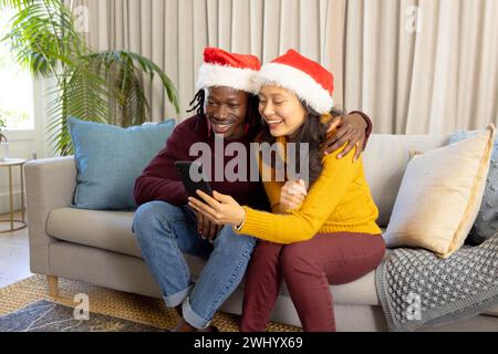 Happy diverse couple in christmas hats having smartphone video call at home, embracing Stock Photo