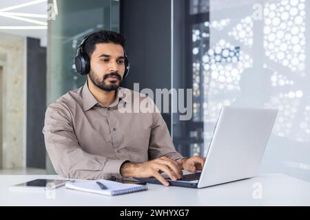 Focused latino male in formal shirt wearing modern headset and using portable computer while working by desk. Contemporary programmer getting ready for video meeting with professional garniture. Stock Photo