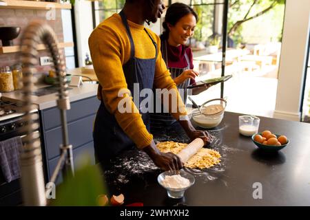 Happy diverse couple baking preparing dough and using tablet in sunny kitchen Stock Photo