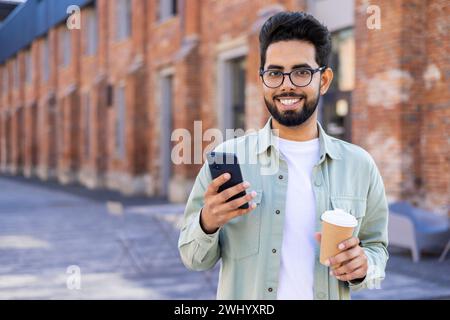 Relaxed middle eastern man holding telephone and plastic coffee cup while walking during daytime. Positive brunet freelancer with flexible work schedule taking break for beverage and scrolling feed. Stock Photo