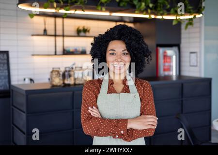 Satisfied black woman in apron standing with crossed arms on chest on background of bar counter. Confident small business owner in service sector ready for accepting clients in beginning of day. Stock Photo