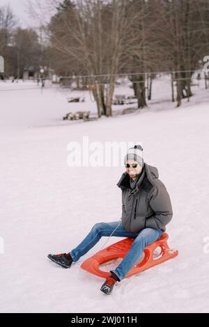 Smiling guy in sunglasses sits on a sled at the snowy edge of the forest Stock Photo