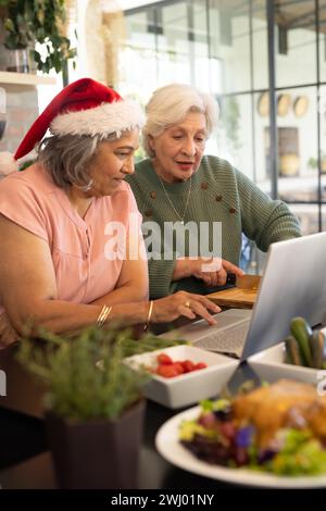 Happy diverse female senior friends in santa hat, using laptop in sunny kitchen at home Stock Photo
