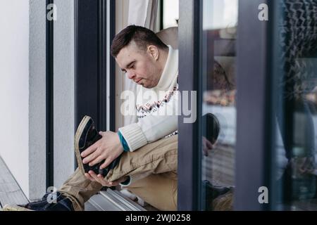 Portrait of young man with Down syndrome putting on shoes, sitting on floor. Stock Photo