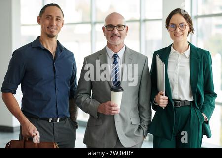 Two young intercultural smiling office managers surrounding confident mature director of business company in grey formalwear Stock Photo