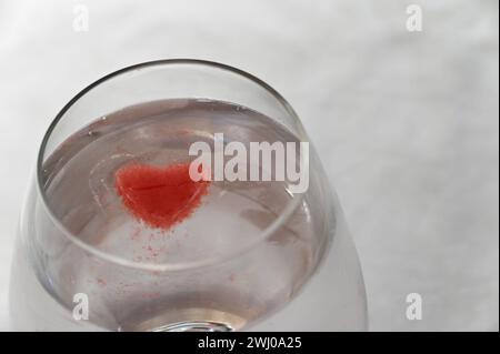 A glass is full of a clear liquid and a bright red, heart shaped ice cube floating in it Stock Photo