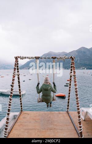 Girl in a jacket and hat swings on a swing on the pier and looks at the sea and mountains. Back view Stock Photo