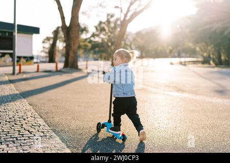 Little girl rides a scooter on an asphalt road and looks away. Back view Stock Photo
