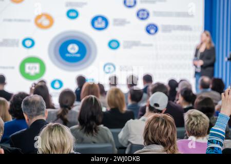 Focused audience listening to a female speaker at a business conference with informational graphics. Stock Photo