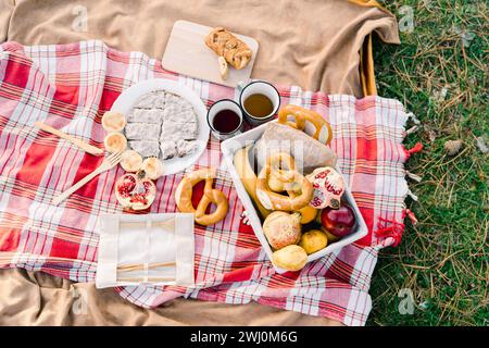 Mugs of coffee stand on a blanket next to buns and fruit in a basket and a sliced pie Stock Photo