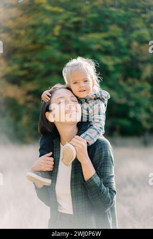 Smiling little girl hugging her mother head while sitting on her shoulders Stock Photo
