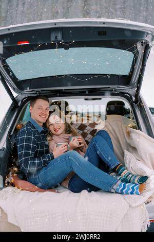 Smiling couple leaning against each other while sitting with mugs of coffee on a blanket in the trunk of a car Stock Photo