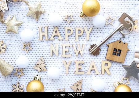 House key with keychain cottage on festive knitted background with stars, lights of garlands. Happy New Year wooden letters, gre Stock Photo