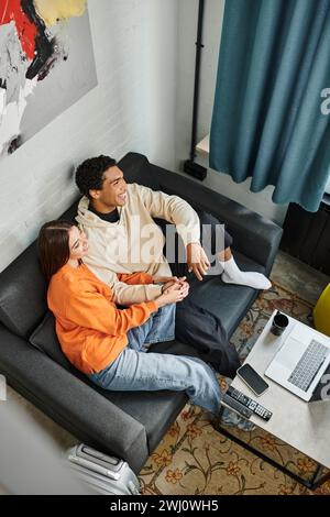 jolly diverse couple enjoys a calm moment while relaxing on sofa with a gadgets and luggage Stock Photo