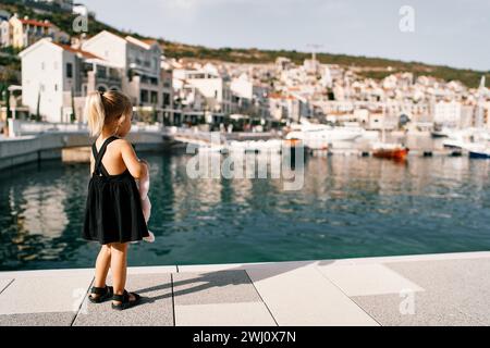 Little girl with a toy in her hands stands on the pier and looks at the marina with yachts. Back view Stock Photo