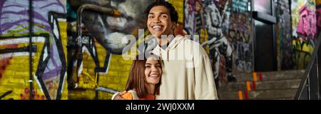 banner of diverse happy couple standing hugging in a graffitied stairwell, sharing a smile Stock Photo