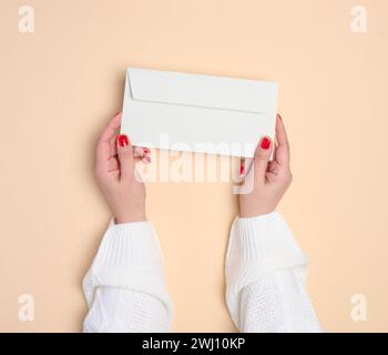 Two female hands holding a white rectangular envelope on a beige background, top view Stock Photo