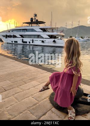 Little girl sits on a bollard on the pier and looks at a moored yacht at sunset Stock Photo