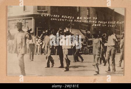 TULSA RACE RIOT(May 31 and June 1, 1921):  A black-and-white photograph of men walking down the middle of a street with hands raised in surrender during the Tulsa Race Massacre in 1921. In the background, a crowd of people line the street in front of a building. Written into the negative of the photograph is [CAPTURED NEGROS ON WAY TO / CONVENTION HALL-DURING TULSA RACE RIOT / JUNE 1ST 1921.]. Stock Photo