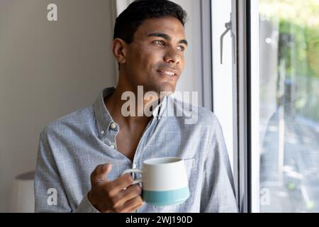 Happy biracial man holding mug looking through window at home Stock Photo