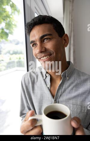 Happy biracial man holding mug looking through window at home Stock Photo