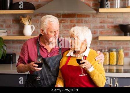 Happy caucasian senior couple cooking, drinking wine and embracing in kitchen Stock Photo