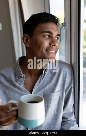 Happy biracial man holding mug looking through window at home Stock Photo