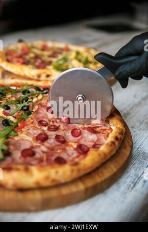 Cutting pizza on slices with special knife. Fresh pizza at background. Hand holding knife. Stock Photo