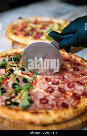 Cutting pizza on slices with special knife. Fresh pizza at background. Hand holding knife. Stock Photo