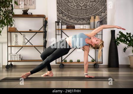 Flexible barefoot lady in activewear practicing Camatkarasana indoors for relaxation during yoga session against shelves potted plants and tapestry on Stock Photo