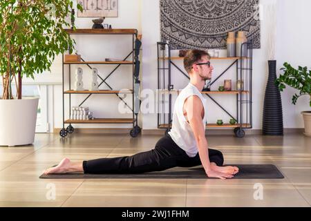 Full body side view of young male in sportswear looking away while sitting on mat in Ardha Kapotasana pose and practicing yoga in cozy room in dayligh Stock Photo