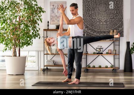 Full body of yoga teacher holding hand of woman while assisting her with Ardha Chandrasana indoors during training against potted green plant shelves Stock Photo