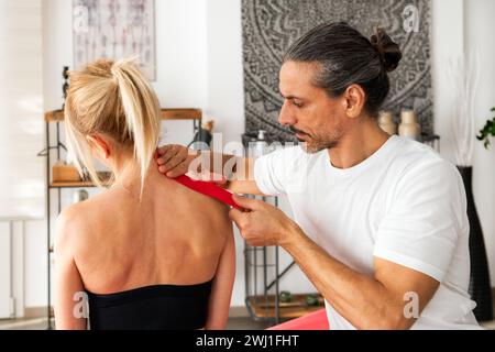 Side view of concentrated adult male therapist with hair bun looking down while sitting applying Kinesio tape on unrecognizable blond female shoulder Stock Photo