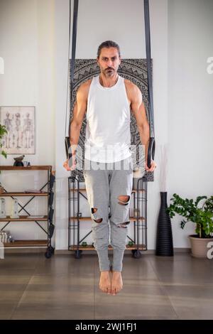 Full body of barefoot confident adult male with beard looking at camera while dipping on gymnastic rings in gym and exercising in daylight Stock Photo