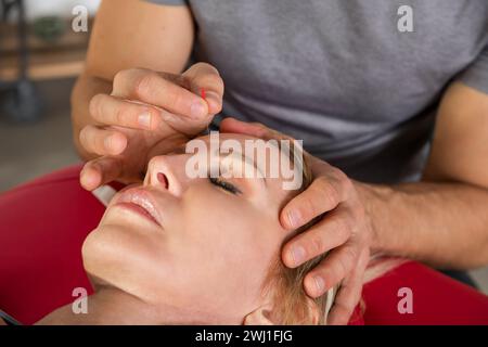 From above crop professional acupuncturist applying needles on woman forehead during acupuncture treatment therapy on face while lying in wellness cen Stock Photo