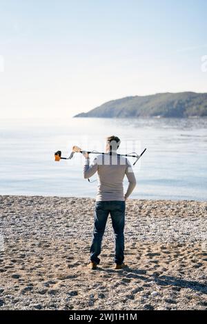 Man with a metal detector on his shoulder stands on the shore and looks ...