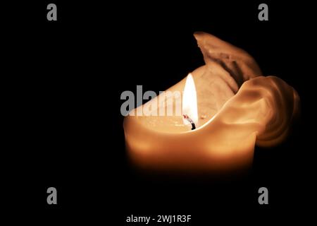 White candle with a flame and melting wax on the edge against a black background, light in the dark for Christmas, advent or New Stock Photo