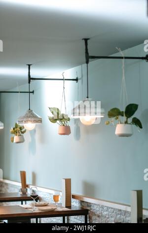 Stylish side view of a bar featuring designer white lamps, hanging plants, dining tables and chairs, with a bar counter in the background. Stock Photo