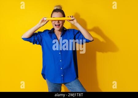 Cheerful woman wearing blue shirt holding giant pencil on yellow background Stock Photo