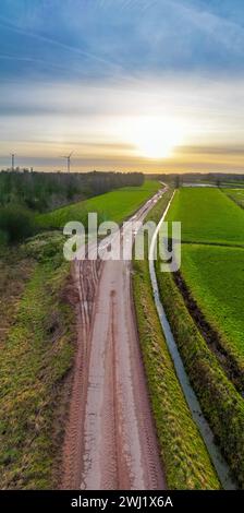 This image is an aerial view capturing the gentle curves of a muddy rural path as it meanders through vibrant green fields. The path leads towards the horizon where the early morning sun bursts with a soft, warm light, suggesting the start of a new day. The contrast between the muddy path and the lush fields is striking, emphasizing the beauty of the rural landscape. In the background, wind turbines align with the rising sun, symbolizing a harmony between agriculture and renewable energy. Aerial Shot of Curvy Rural Path at Dawn. High quality photo Stock Photo