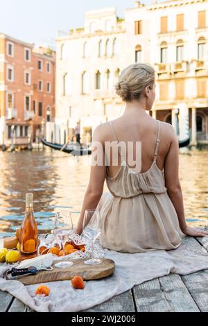 Lovely young woman picturesque picnic on the wooden gondola dock with rose wine, fruits and snack on wooden pier Stock Photo