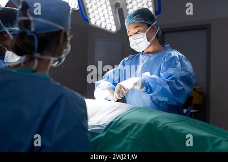 Diverse female doctor with face mask doing surgery in group of doctors in hospital operating room Stock Photo