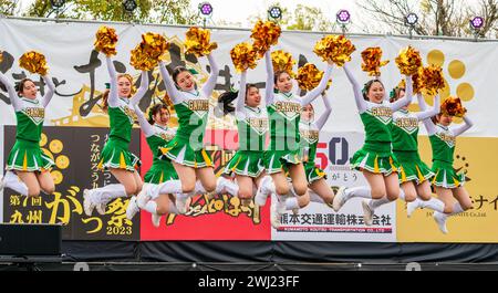 Japanese teenage women cheerleader Yosakoi dance team in green costumes dancing on stage holding gold glittery pom poms. All in mid air after jumping. Stock Photo