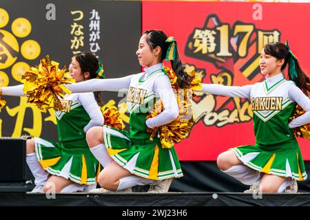 Japanese teenage women cheerleader Yosakoi dance team in green costumes dancing on stage holding gold glittery pom poms. All kneeling on stage. Stock Photo