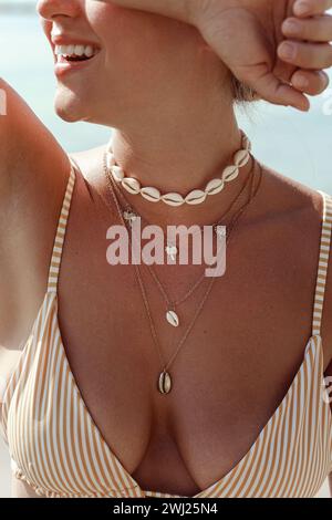 Cheerful woman in bikini wearing golden necklace with palm trees and seashells on a sunlit beach Stock Photo