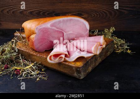 Traditional South Tyrolean farmer's ham sliced and in one piece offered with dried herbs as a close-up on a rustic cutting board Stock Photo